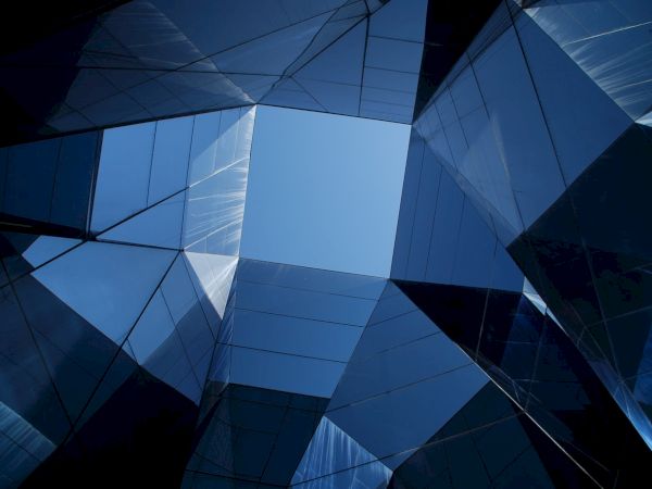 A geometric view looking up at a blue sky through modern glass skyscrapers, forming angular reflections and lines.