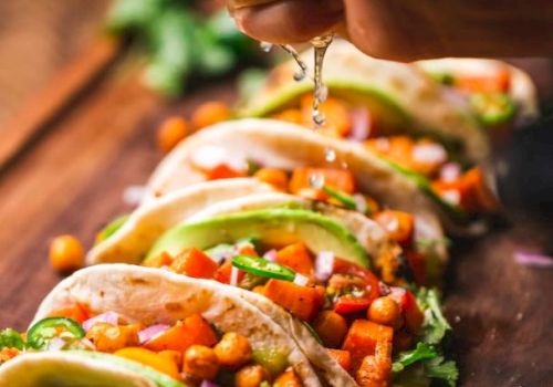 The image shows a person squeezing lime over a row of colorful tacos filled with chickpeas and vegetables, garnished with avocado slices.