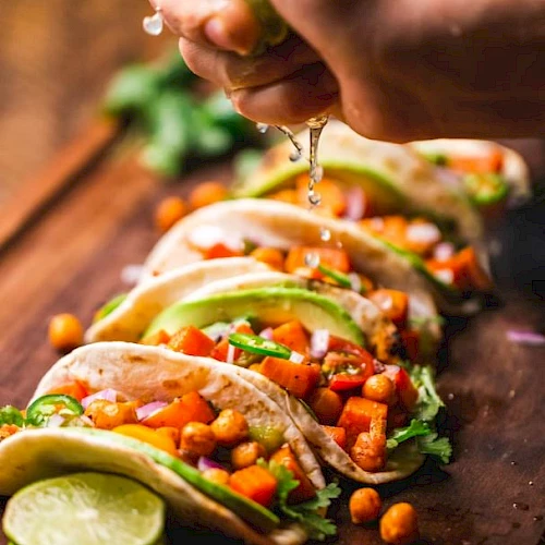 The image shows a person squeezing lime over a row of colorful tacos filled with chickpeas and vegetables, garnished with avocado slices.