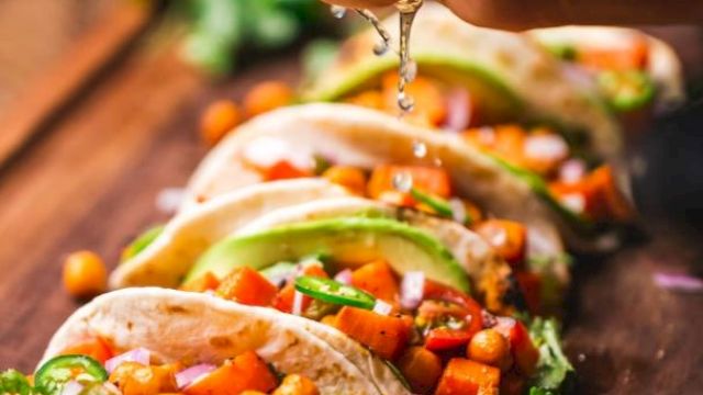 The image shows a person squeezing lime over a row of colorful tacos filled with chickpeas and vegetables, garnished with avocado slices.