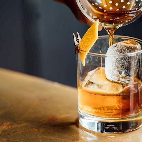 A cocktail is being poured into a glass with ice and garnished with a lemon twist on a wooden surface.