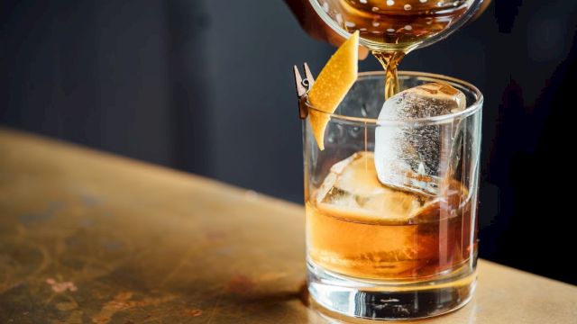 A cocktail is being poured into a glass with ice and garnished with a lemon twist on a wooden surface.
