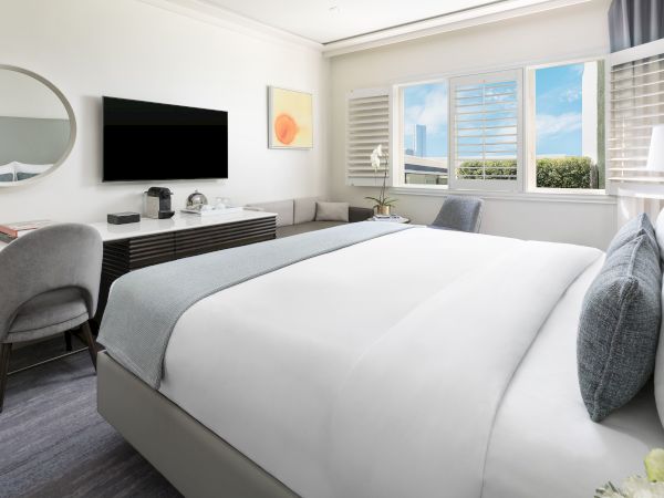 A modern hotel room with a large bed, desk, TV, and a view through a window. There's a circular mirror and minimal decor.