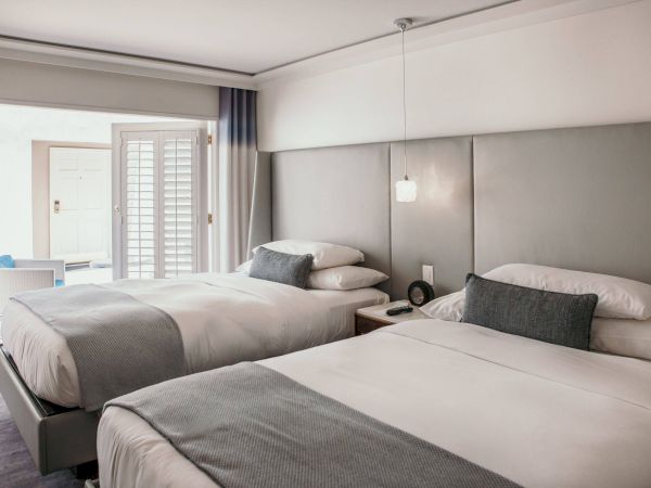 The image shows a hotel room with two neatly made beds, gray accents, a bedside table, a lamp, and an open door leading to a patio.