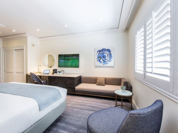 The image shows a modern hotel room with a bed, TV, sofa, chair, and table, decorated in neutral tones with a piece of abstract art.