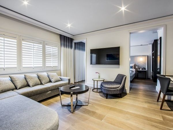 A modern living room with a gray sofa, round tables, TV, and visible bedroom, featuring light wood flooring and ceiling lights.