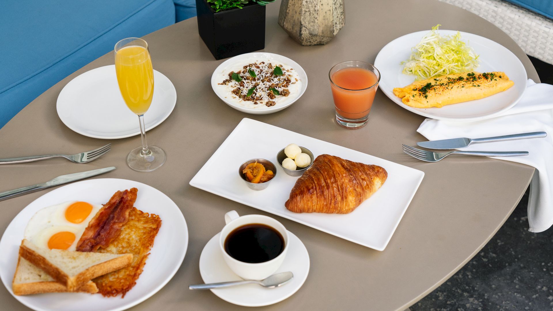 A breakfast spread with eggs, bacon, croissant, coffee, juice, omelette, and a savory dish, set on a round table with silverware and plants.