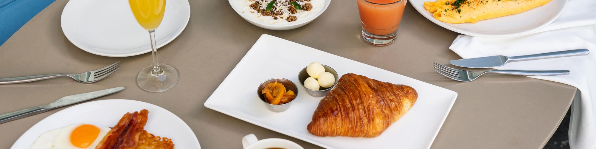 A breakfast spread with eggs, bacon, croissant, coffee, juice, omelette, and a savory dish, set on a round table with silverware and plants.