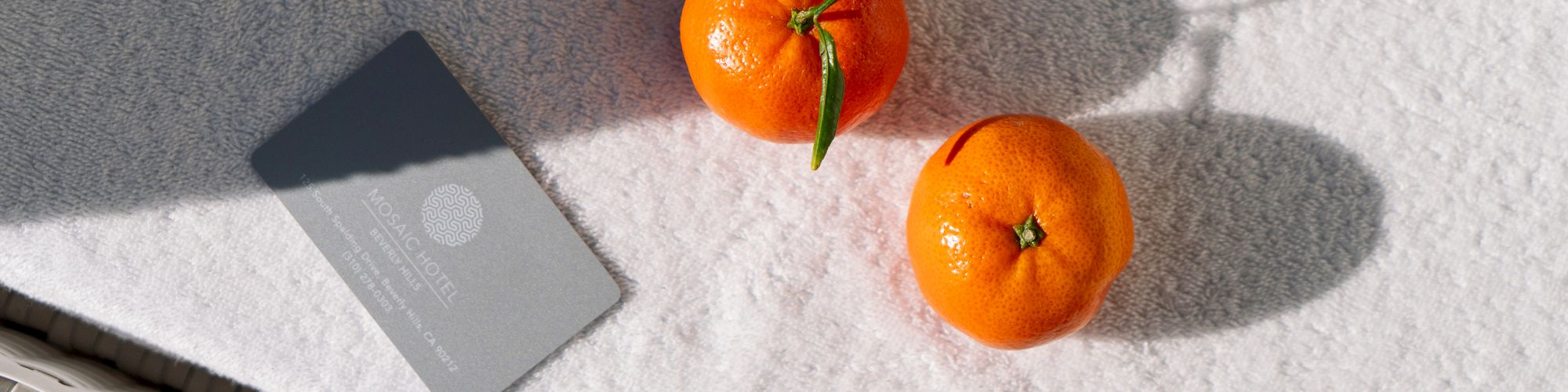 Two oranges with leaves sit next to a hotel card on a textured white surface in the sunlight.