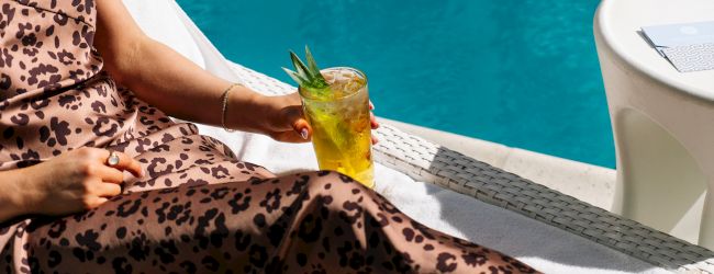 A person in a patterned dress relaxes by a pool, holding a tropical drink with pineapple garnish.