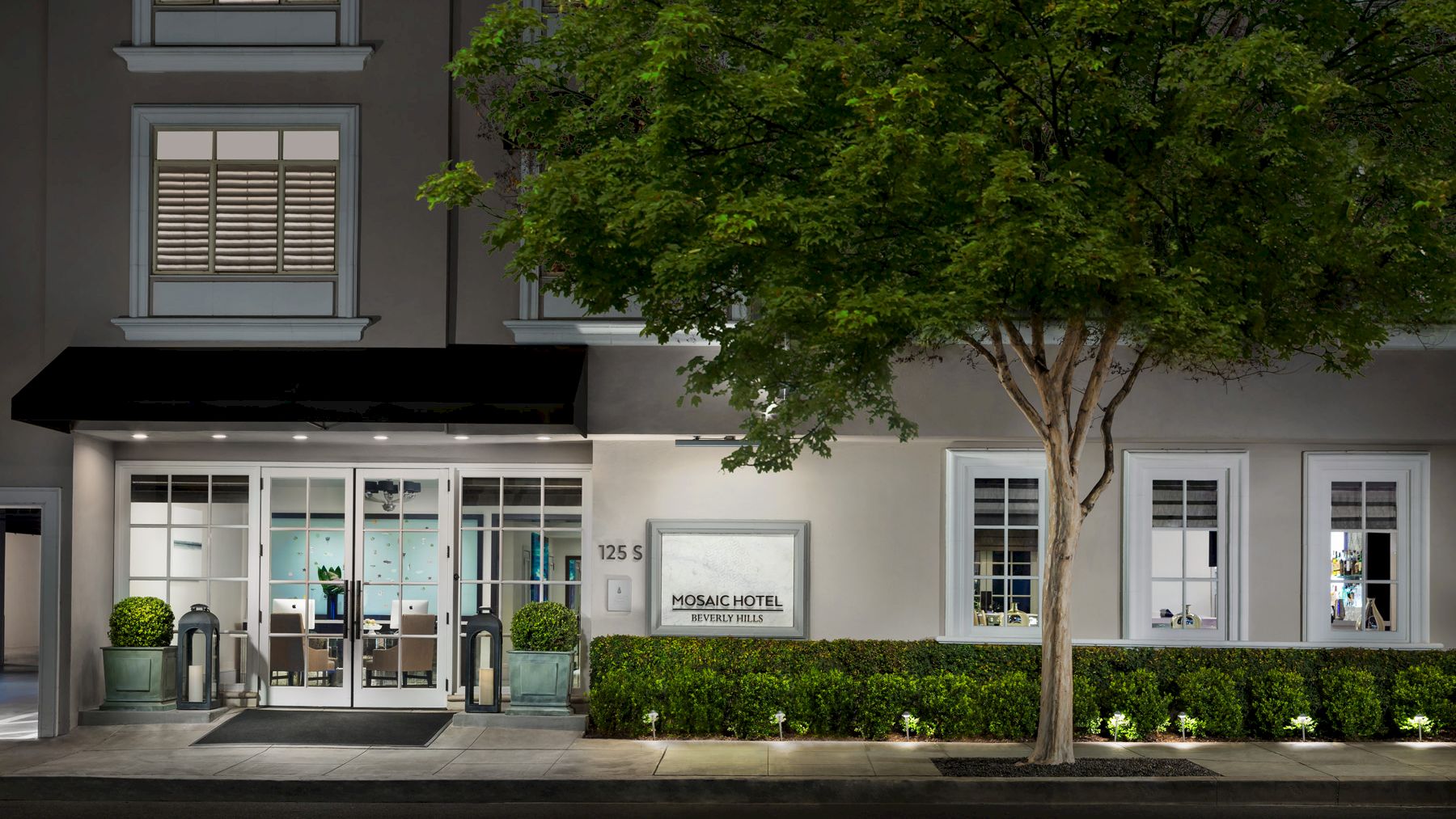 A building exterior with a glass entrance, potted plants, and a tree on the right side, lit by streetlights, creating a welcoming ambiance.