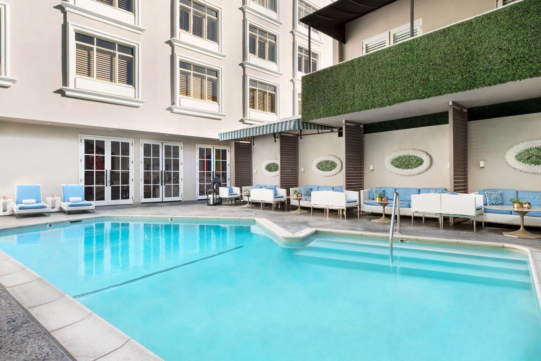 A swimming pool with light blue water is surrounded by seating areas and a building with large windows and greenery.