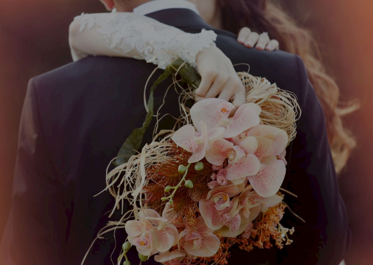 A person is embracing someone, holding a bouquet of pink and white flowers with greenery, in a romantic setting.