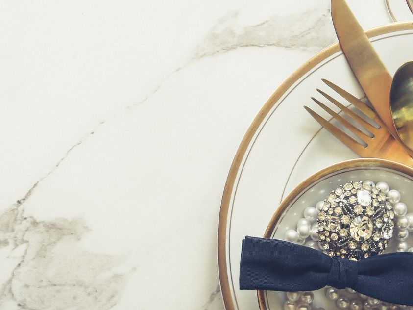 Elegant table setting with gold cutlery, pearl decorations, and a blue bow tie, all arranged on white plates atop a marble surface.