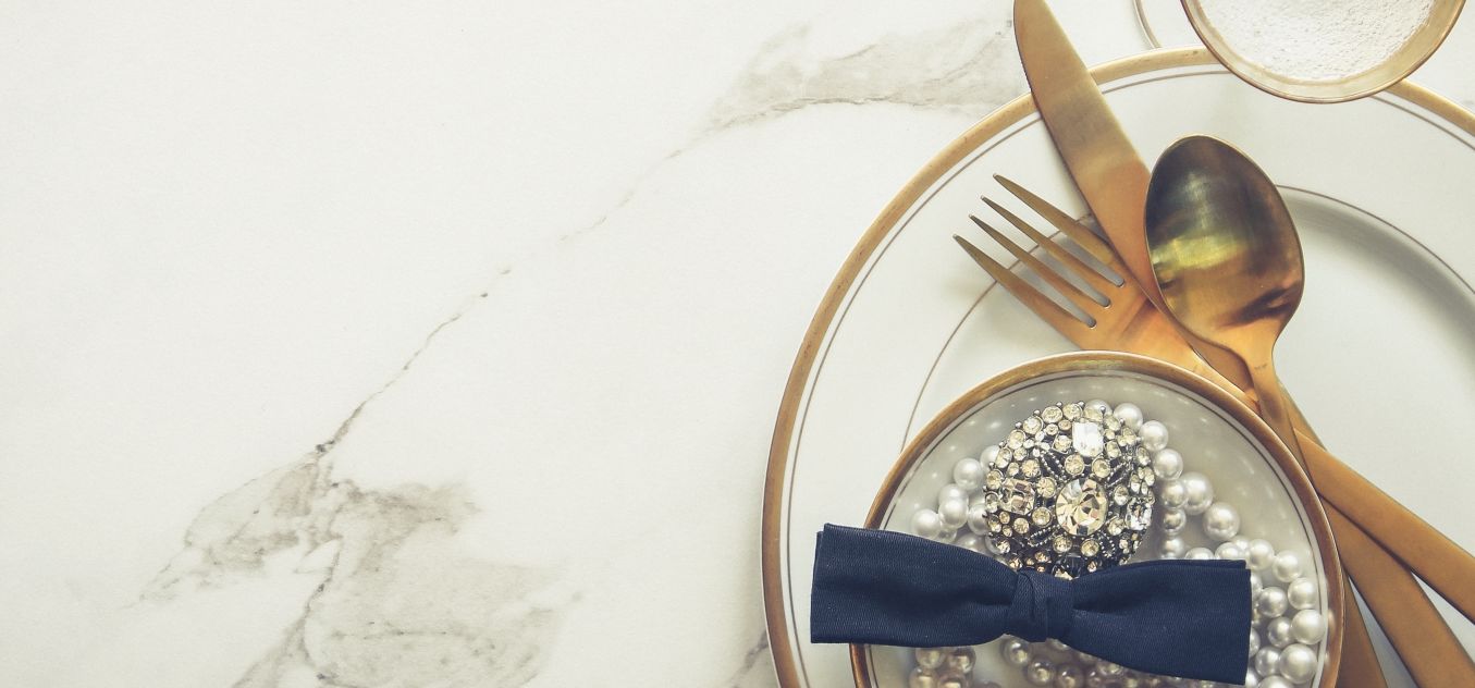 Elegant table setting with gold cutlery, pearl decorations, and a blue bow tie, all arranged on white plates atop a marble surface.