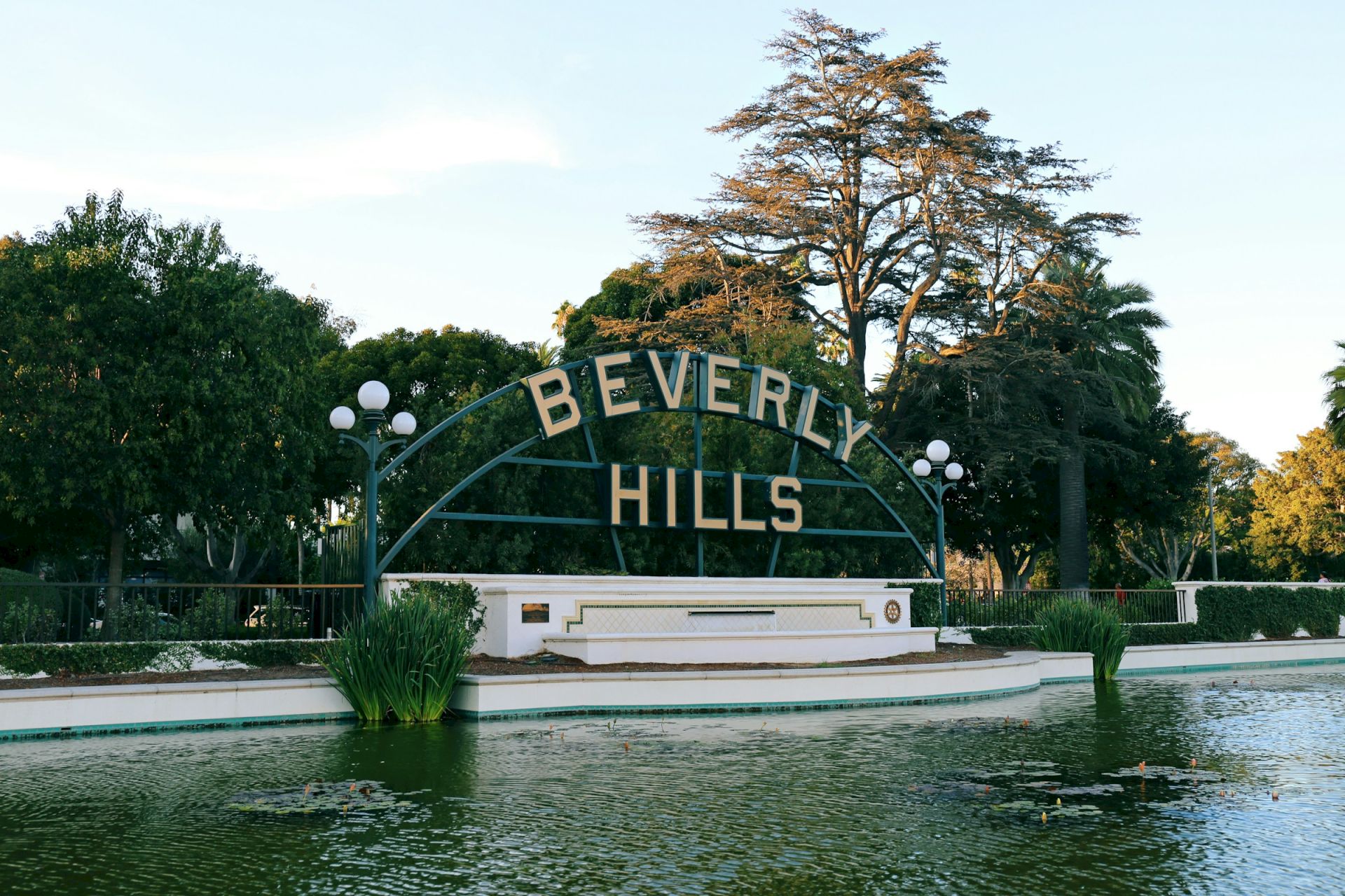 The image shows the iconic "Beverly Hills" sign above a pond, surrounded by trees and lamps, under a clear blue sky.