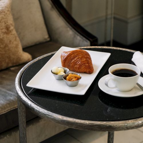 A croissant with butter and jam on a tray, paired with a coffee cup, napkin, on a table by a sofa with a cushion.