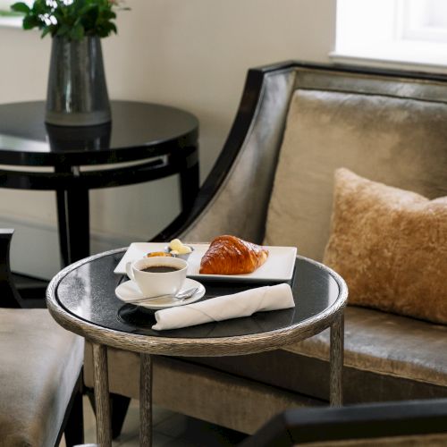 A cozy seating area with a small table featuring a cup of coffee and a croissant, alongside a cushioned chair and a vase of flowers.