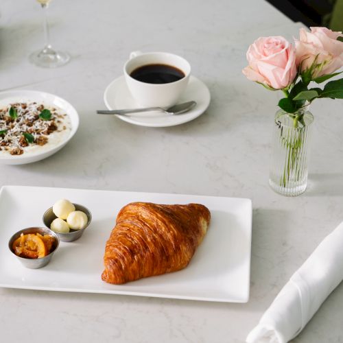A breakfast setup with a croissant, butter, jam, coffee, yogurt, and a mimosa. There's a vase with pink roses on the table.