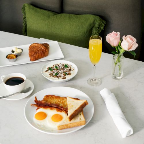 A breakfast spread with eggs, toast, bacon, croissant, coffee, mimosa, fruit, butter, and roses on a table.