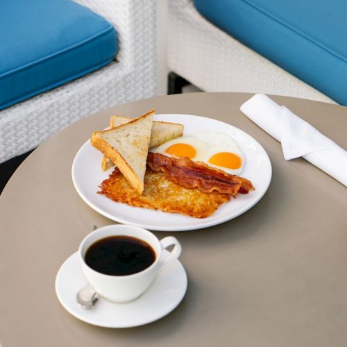 A breakfast plate with eggs, bacon, hash browns, and toast is on a table next to a cup of coffee and a napkin.