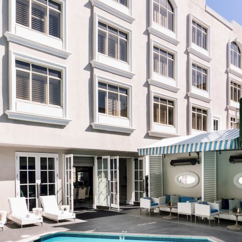 A swimming pool is surrounded by lounge chairs, next to a multi-story building with large windows and striped awnings.