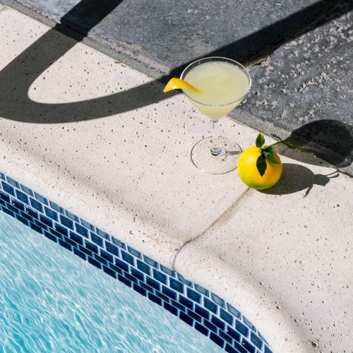 A lemon cocktail and a lemon are placed by a poolside on a sunny day, casting a shadow on the stone pavement.