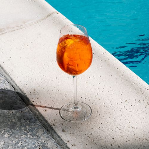 A glass of orange beverage sits by the edge of a pool, casting a shadow on the concrete surface.