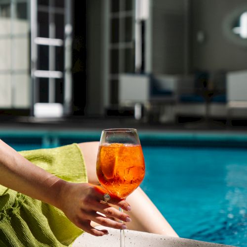 A person relaxes by a pool, holding a glass of orange drink, with a green towel over their lap.