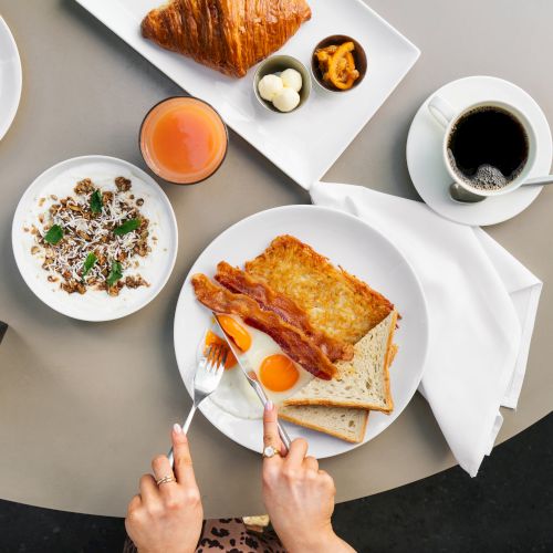 A breakfast spread with toast, bacon, eggs, granola, croissant, juice, and coffee on a table. Hands holding utensils.