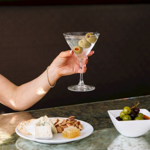A person holds a martini with olives over a table with cheese, crackers, and a bowl of olives.