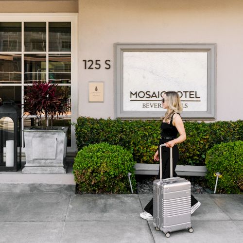 A person with a suitcase walks past Mosaic Hotel in Beverly Hills, located at 125 S, with plants outside.