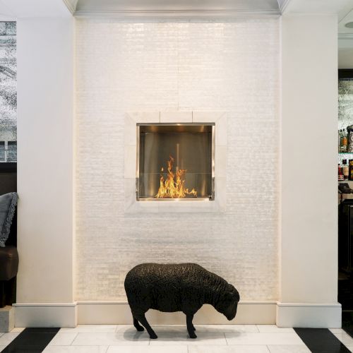 A black sheep sculpture on a white tile floor, in front of a modern wall-mounted fireplace set in a white brick wall.