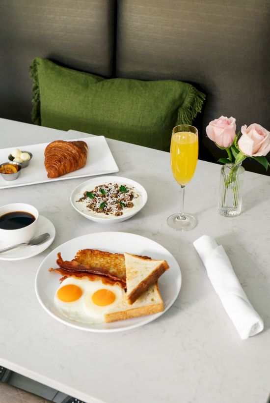 A breakfast spread with eggs, bacon, toast, coffee, a croissant, a dessert, a drink, and roses on a table.
