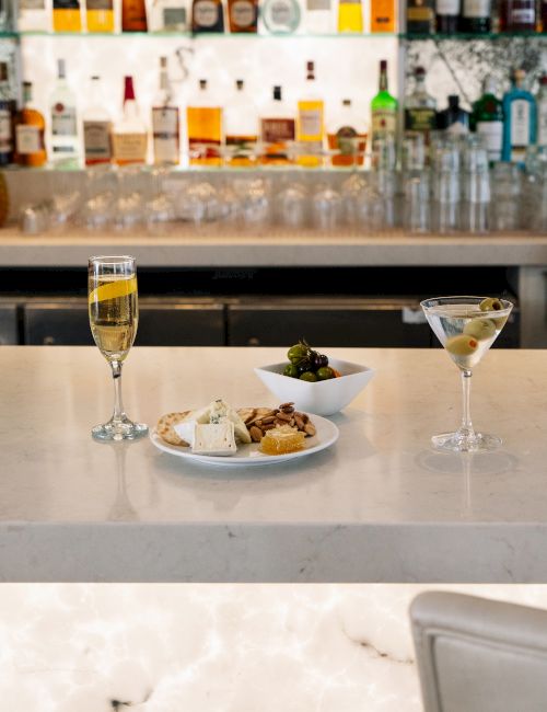 A bar setup with two drinks, a plate of food, and a bowl of olives on a counter, with bottles visible in the background.