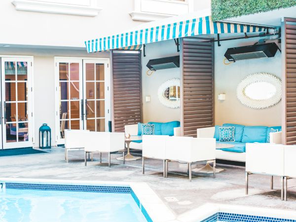 Poolside lounge area with white furniture, blue cushions, and striped awning; adjacent to a building with glass doors.