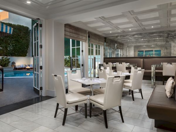 A modern dining area with white chairs opens to a poolside patio, featuring comfortable outdoor seating and soft lighting for ambiance.