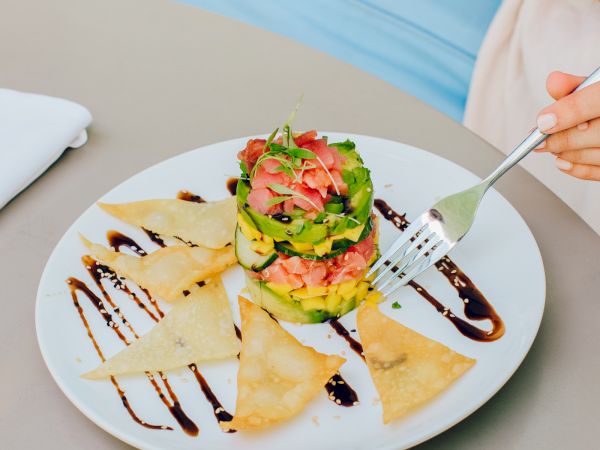 A person is about to eat a colorful dish with avocado, tomato, and wonton chips drizzled with sauce on a round white plate.
