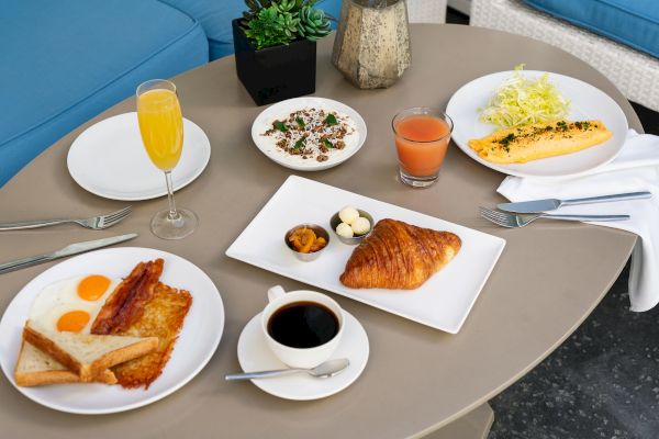Breakfast spread with sunny-side-up eggs, bacon, toast, croissant, fruit cups, coffee, juice, salad, and yogurt on a round table.