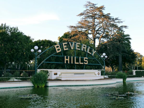 The image shows the Beverly Hills sign in a landscaped area with water and trees in the background.
