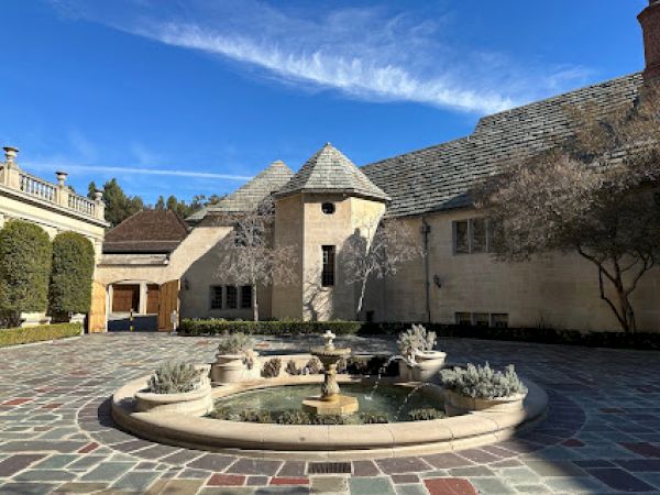 A courtyard with a round fountain in the center, surrounded by stone buildings and a clear blue sky above, creating a serene setting.