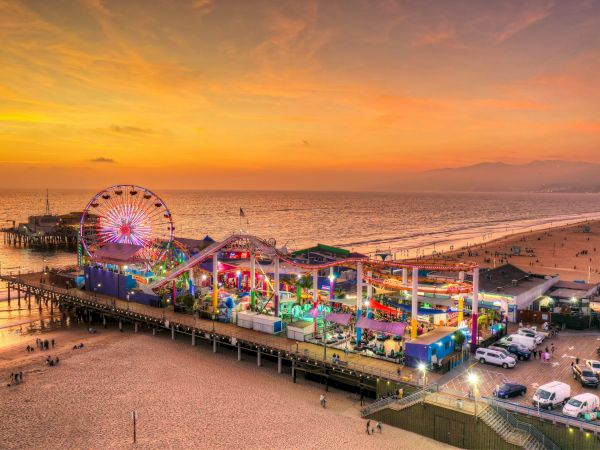 Amusement park on a pier at sunset, featuring a Ferris wheel, roller coaster, and vibrant lights, with a beach and ocean in the background.