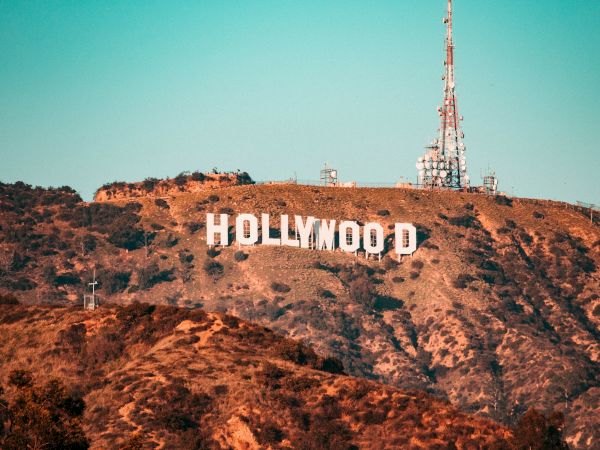The image shows the iconic Hollywood Sign on a hillside with communication towers, set against a clear blue sky.