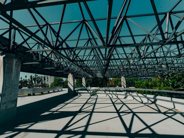 The image shows an architectural structure with metal beams casting geometric shadows on the ground, amidst a backdrop of greenery and sky.
