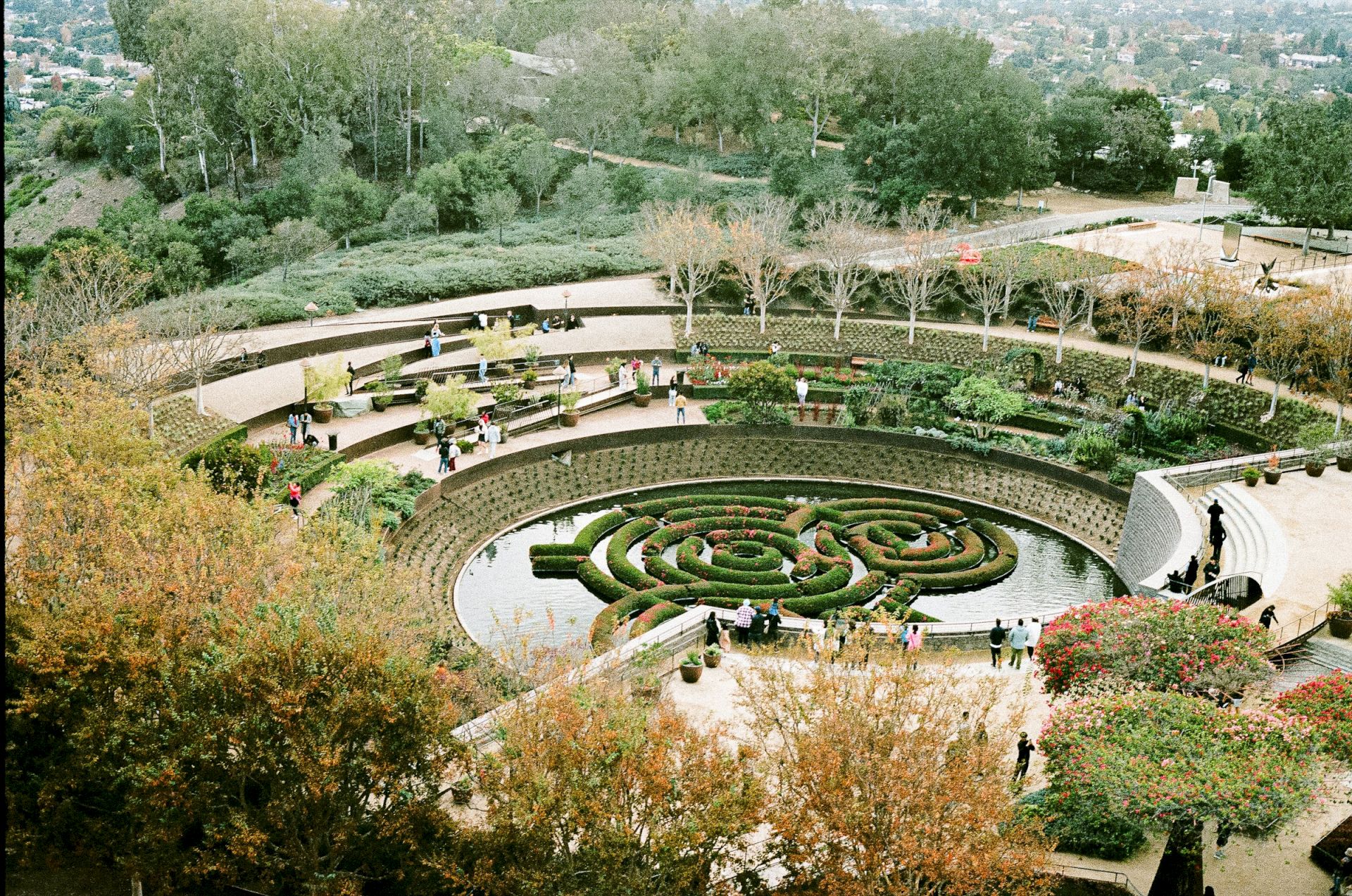 An aerial view of a landscaped garden featuring circular paths, a maze-like design around a pond, and surrounded by trees and visitors.