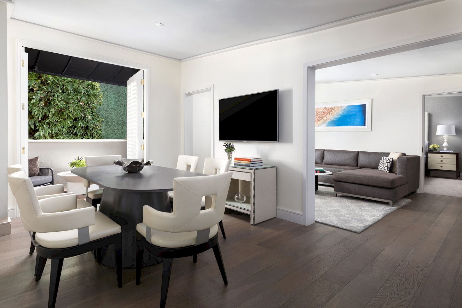 A modern dining room with a dark table and white chairs, opening to a living room with a sofa, art piece, and wall-mounted TV.