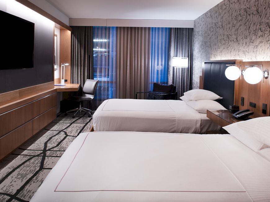 The image shows a modern hotel room with two beds, a wall-mounted TV, a desk by the window, and contemporary decor and lighting.