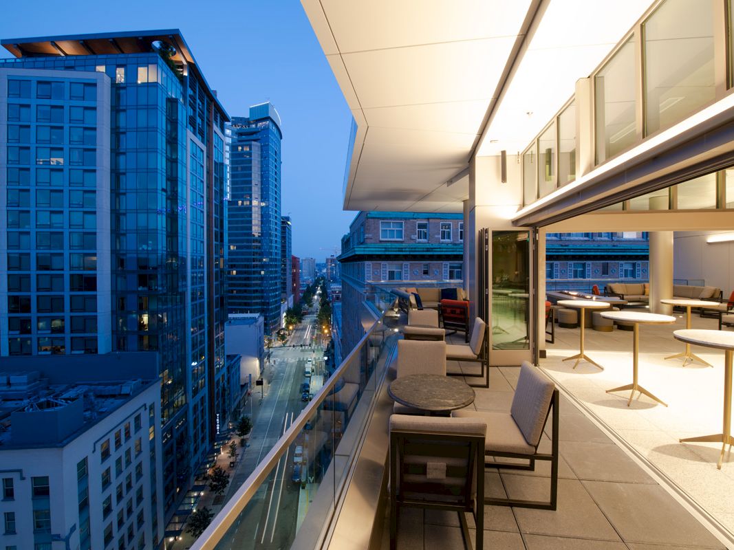 A modern rooftop terrace with seating overlooks a cityscape with tall glass buildings and a street below.