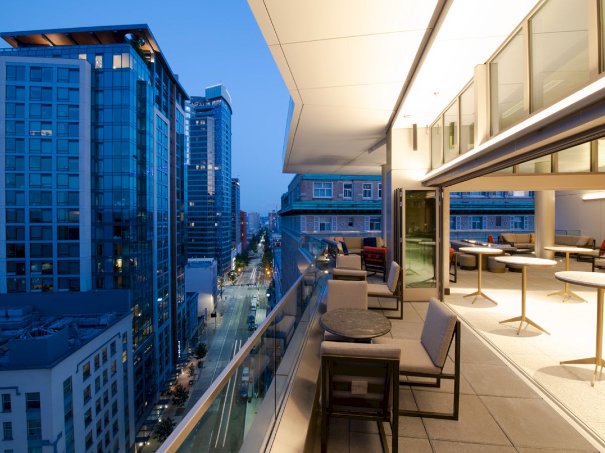 The image shows a modern cityscape from a balcony at dusk, featuring buildings, tables, chairs, and indoor-outdoor dining spaces.