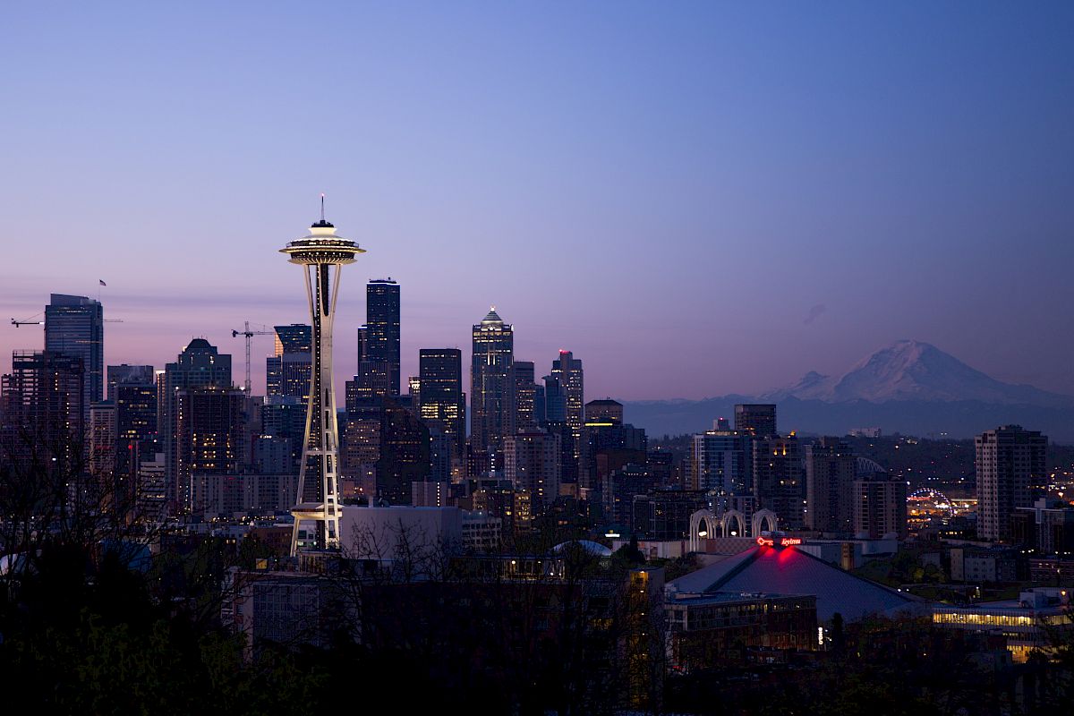 A city skyline at dusk with the Space Needle, other buildings, and a distant mountain under a twilight sky.